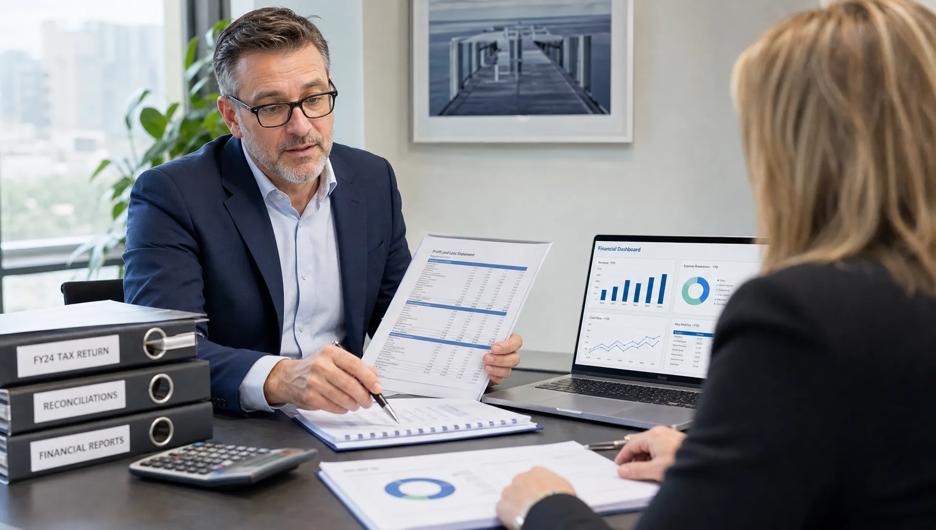 A professional Australian accountant and business director reviewing tax documents, reconciled accounts and financial dashboards on a meeting table, with organised folders, a calculator and a laptop screen facing the participants.
