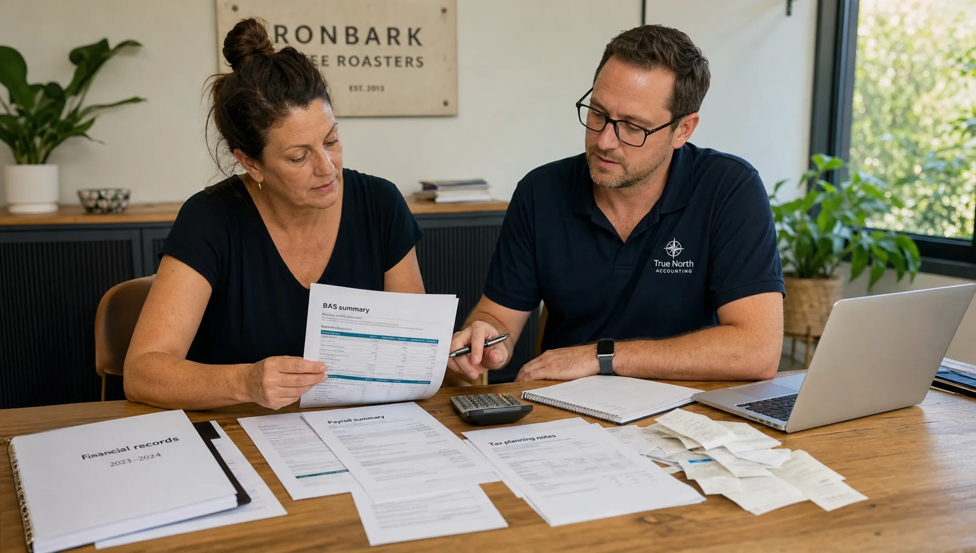 An Australian small business owner and accountant review BAS summaries, payroll reports, receipts and tax planning notes at a meeting table.